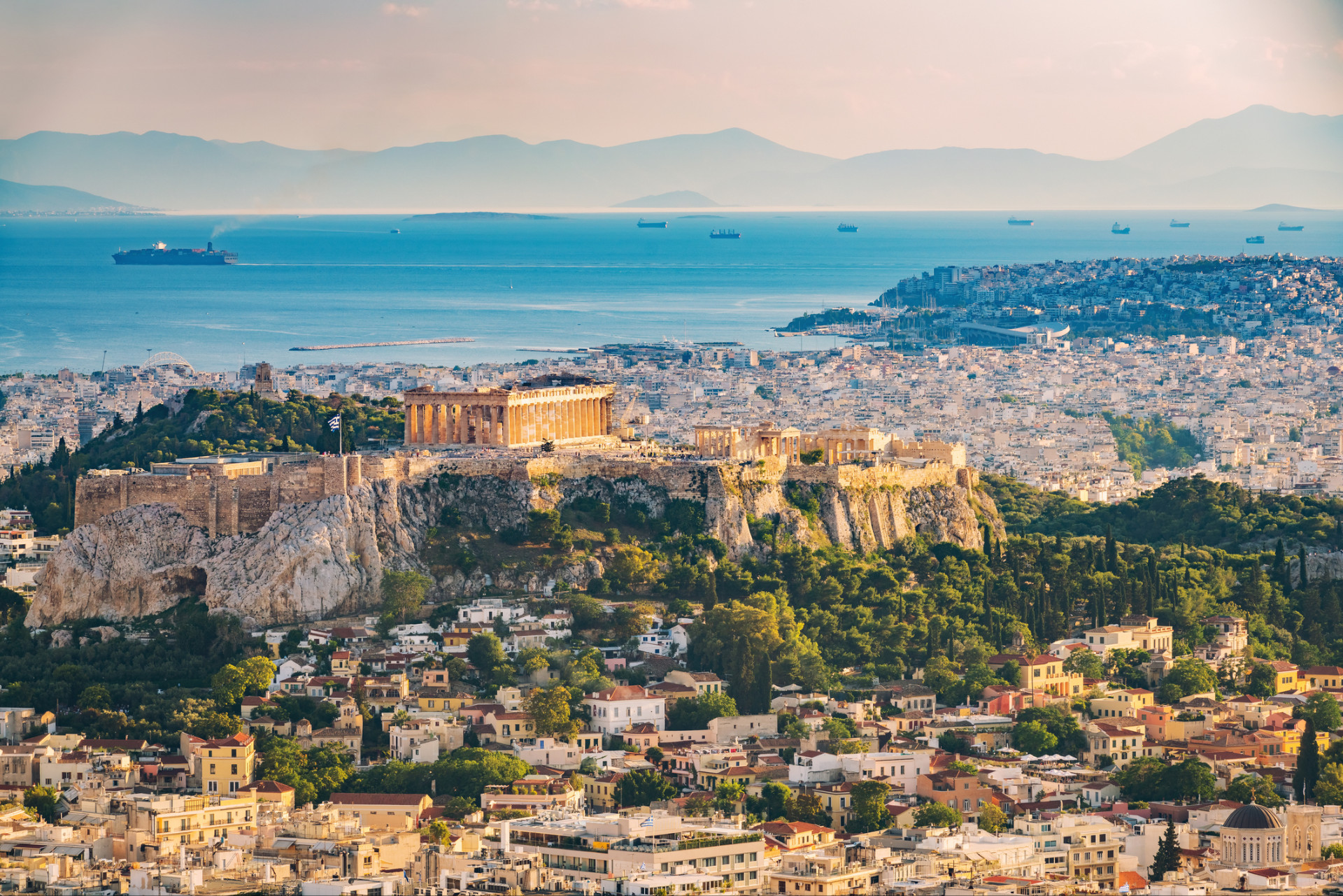 Sun-lit view of the Acropolis from above, the historical centre and neighbourhoods of Athens, with the sea in the background.